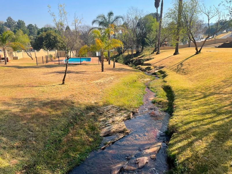 Expansive communal garden area featuring a natural stream, landscaped lawns, and a fenced swimming pool facility.