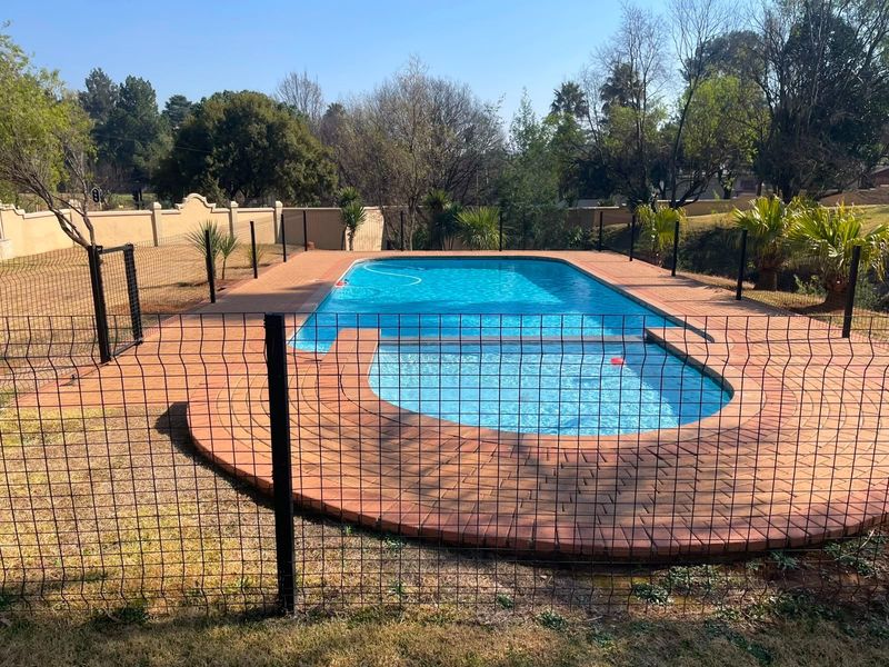 A well-maintained outdoor swimming pool area featuring brick paving, secure perimeter fencing, and a boundary wall in a landscaped garden setting.