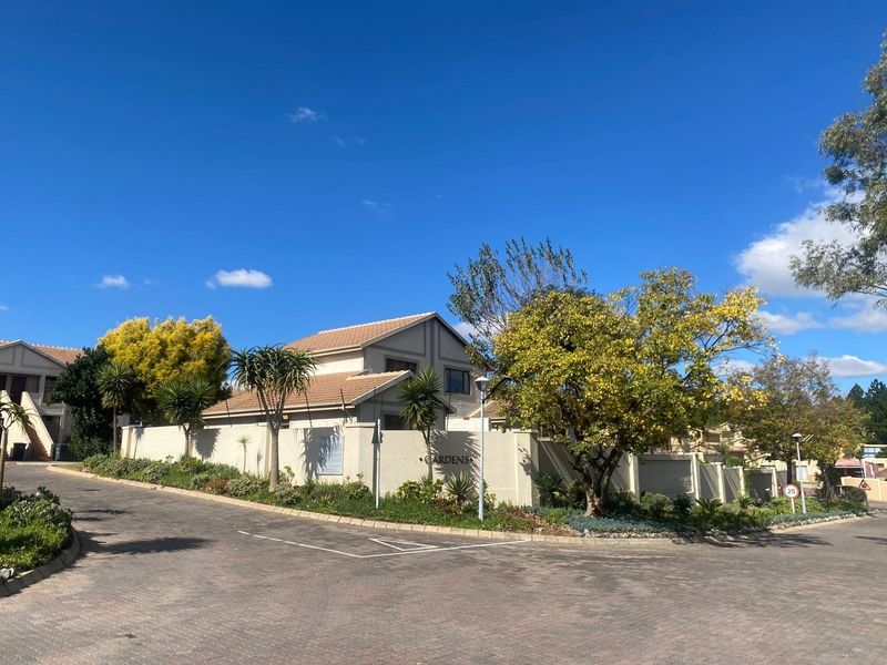 Street view of the townhouse complex entrance featuring paved driveway, perimeter walling, and established landscaping under a clear blue sky.