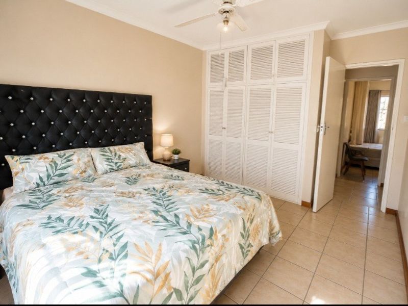 A bedroom featuring tiled flooring, built-in louvered cupboards, and a ceiling fan, with an open doorway leading to an adjacent room.