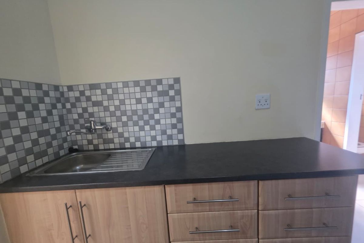 A compact kitchen area featuring light wood-finish cabinetry, a dark laminate countertop, a stainless steel sink, and a modern checkered tile splashback.