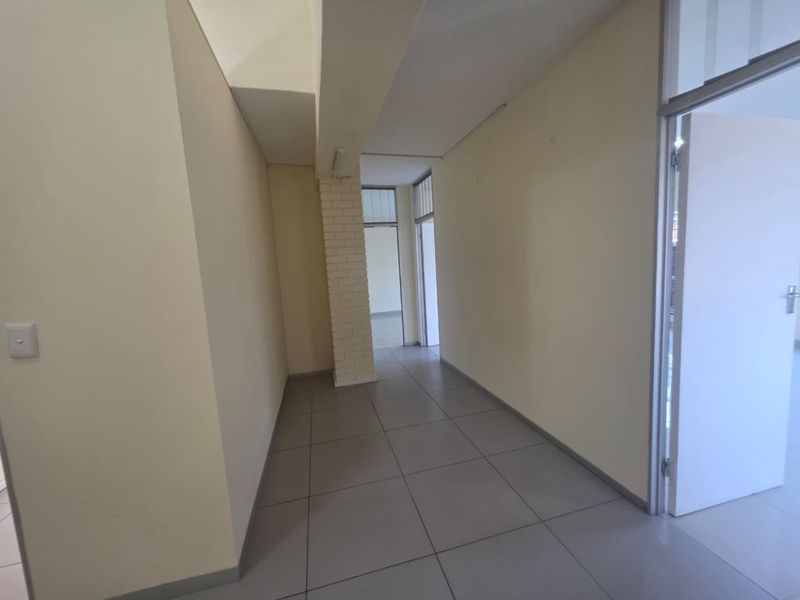 A clean, empty commercial hallway featuring neutral tiled flooring, an exposed brick support column, and multiple access points with aluminium door frames.