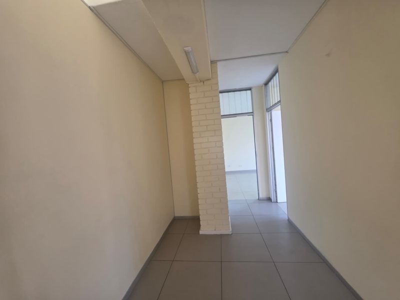 A clean, empty commercial hallway featuring tiled flooring, a central exposed brick column, and functional drop ceiling lighting.