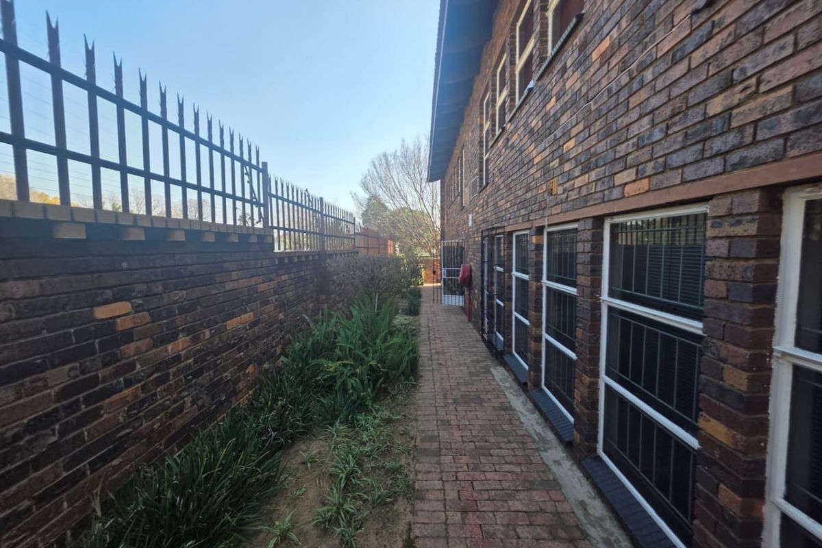 A side exterior view of a commercial brick building featuring a paved walkway, security fencing, and window security bars.