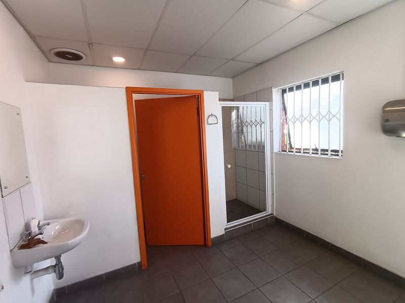 A functional industrial-style bathroom featuring a wall-mounted basin, tiled flooring, a shower enclosure, and a suspended ceiling with integrated lighting.