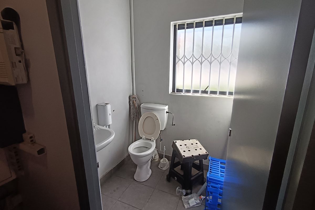 A functional industrial-style bathroom featuring a standard toilet, wall-mounted washbasin, and a window fitted with security bars.