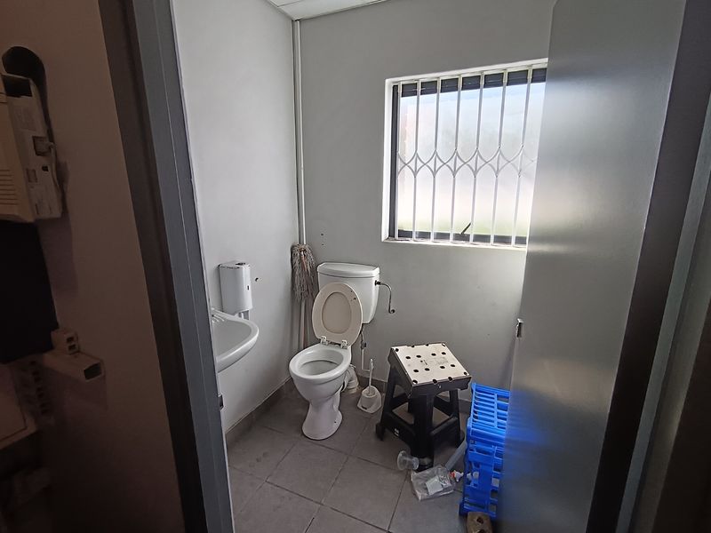 A functional industrial-style bathroom featuring a standard toilet, wall-mounted washbasin, and a window fitted with security bars.