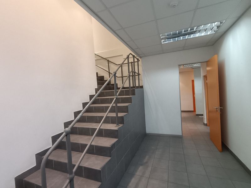 A clean, functional industrial hallway featuring a tiled staircase with metal handrails, suspended ceiling, and neutral finishes.