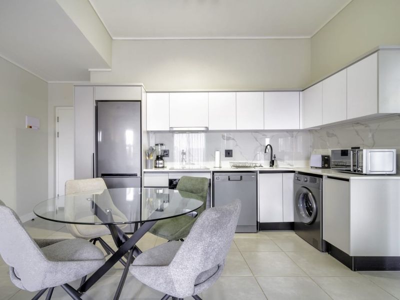 Modern open-plan kitchen featuring sleek white cabinetry, marble-look splashback, integrated appliances, and tiled flooring, seamlessly flowing into the dining area.