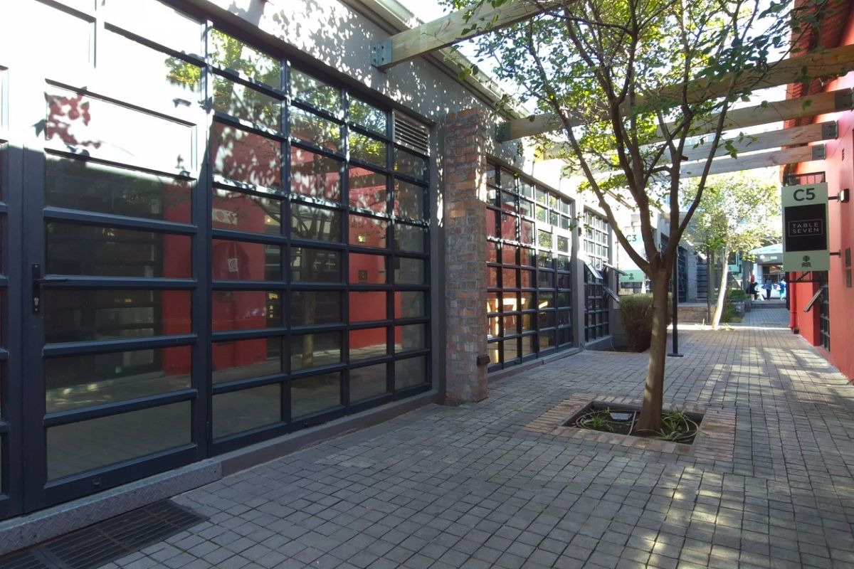 A well-maintained outdoor walkway featuring industrial-style glass doors, brick paving, and a timber pergola structure in a commercial retail setting.