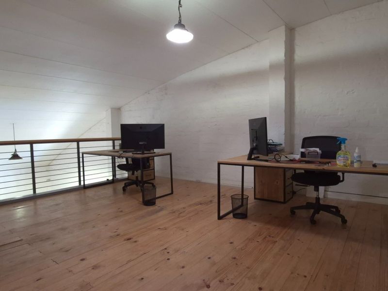 An open-plan mezzanine office space featuring timber flooring, exposed brick walls, and a metal balustrade overlooking the lower level.