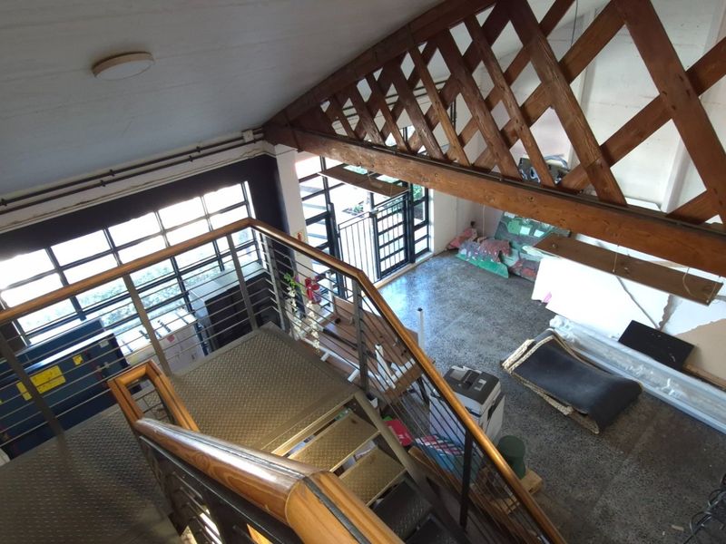 An elevated view of an industrial-style open plan space featuring a metal staircase, exposed timber trusses, and large windows with concrete flooring.