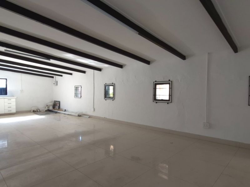 Spacious open-plan retail or office area featuring exposed ceiling beams, tiled flooring, and a small kitchenette corner with security-barred windows.