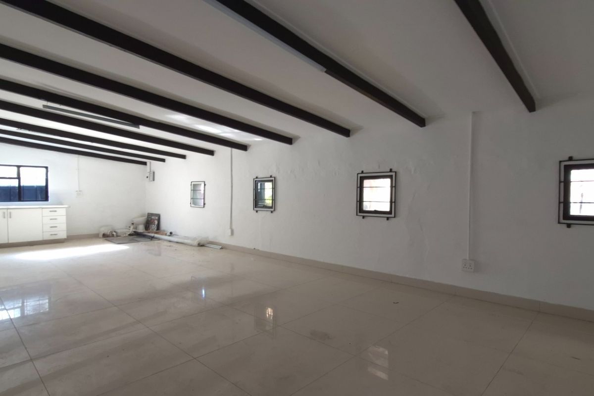 Spacious open-plan retail or office area featuring exposed ceiling beams, tiled flooring, and a small kitchenette corner with security-barred windows.
