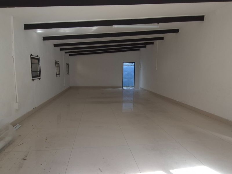 A spacious, empty open-plan retail area featuring tiled flooring, exposed ceiling beams, and multiple windows with security bars.