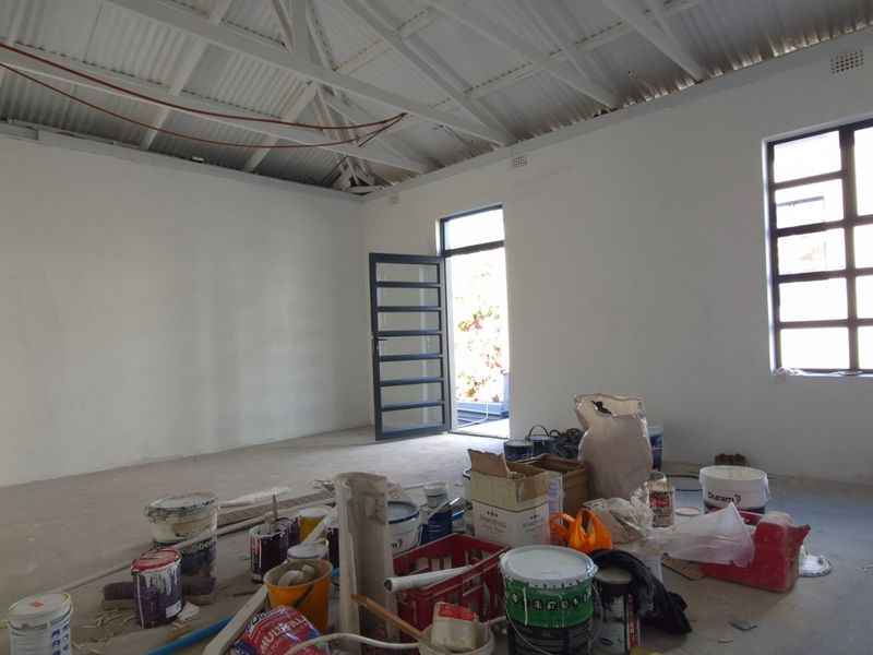 An open-plan retail space currently under renovation, featuring exposed roof trusses, white walls, and steel-framed windows and doors.