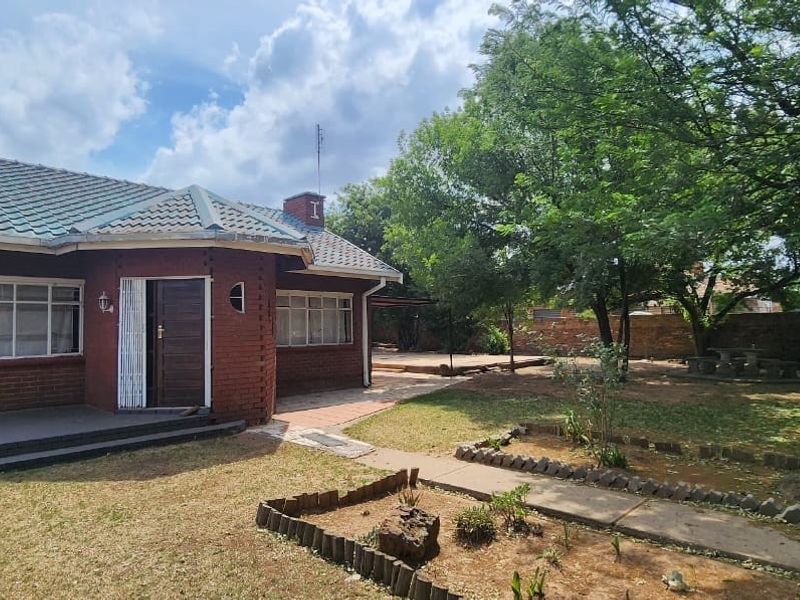Rear view of a brick house featuring a tiled roof, chimney, and a paved patio area leading into a garden with mature trees.