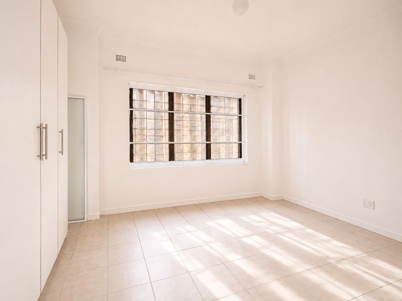 A bright, empty bedroom featuring neutral tiled flooring, built-in cupboards, and a window fitted with security bars.