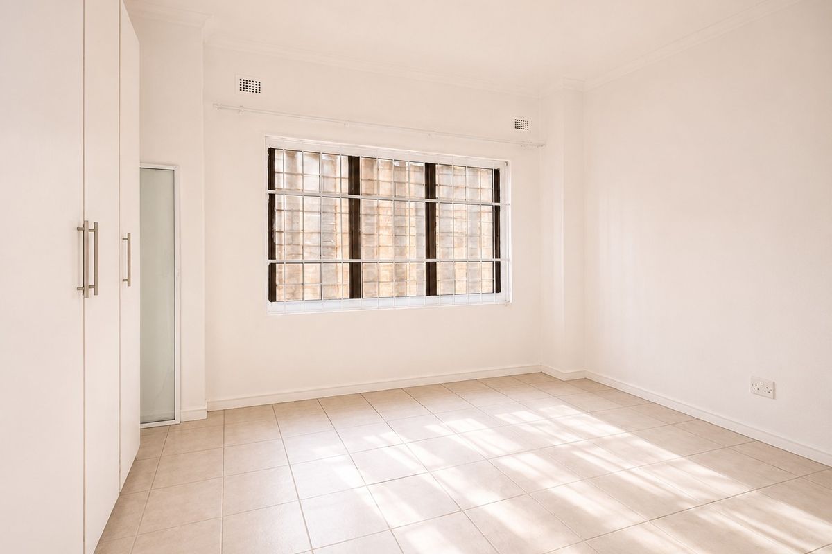A bright, empty bedroom featuring neutral tiled flooring, built-in cupboards, and a window fitted with security bars.