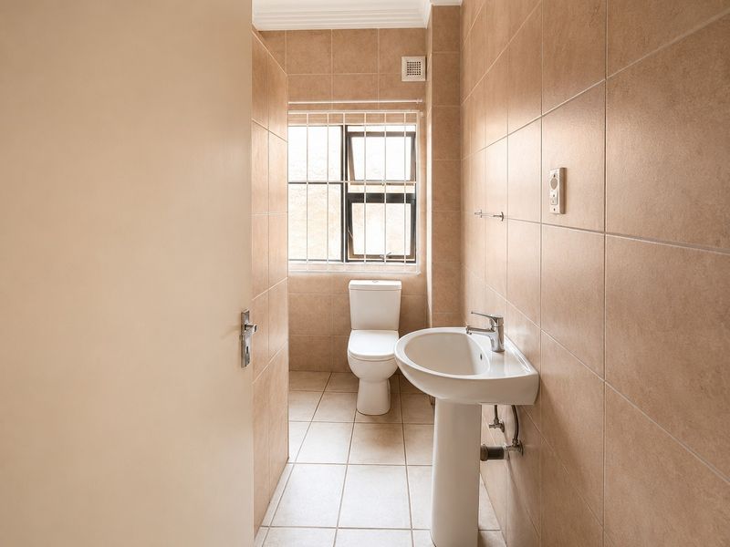 A clean, functional bathroom featuring neutral floor-to-ceiling wall tiles, a pedestal basin, and a toilet, with natural light from a window with security bars.