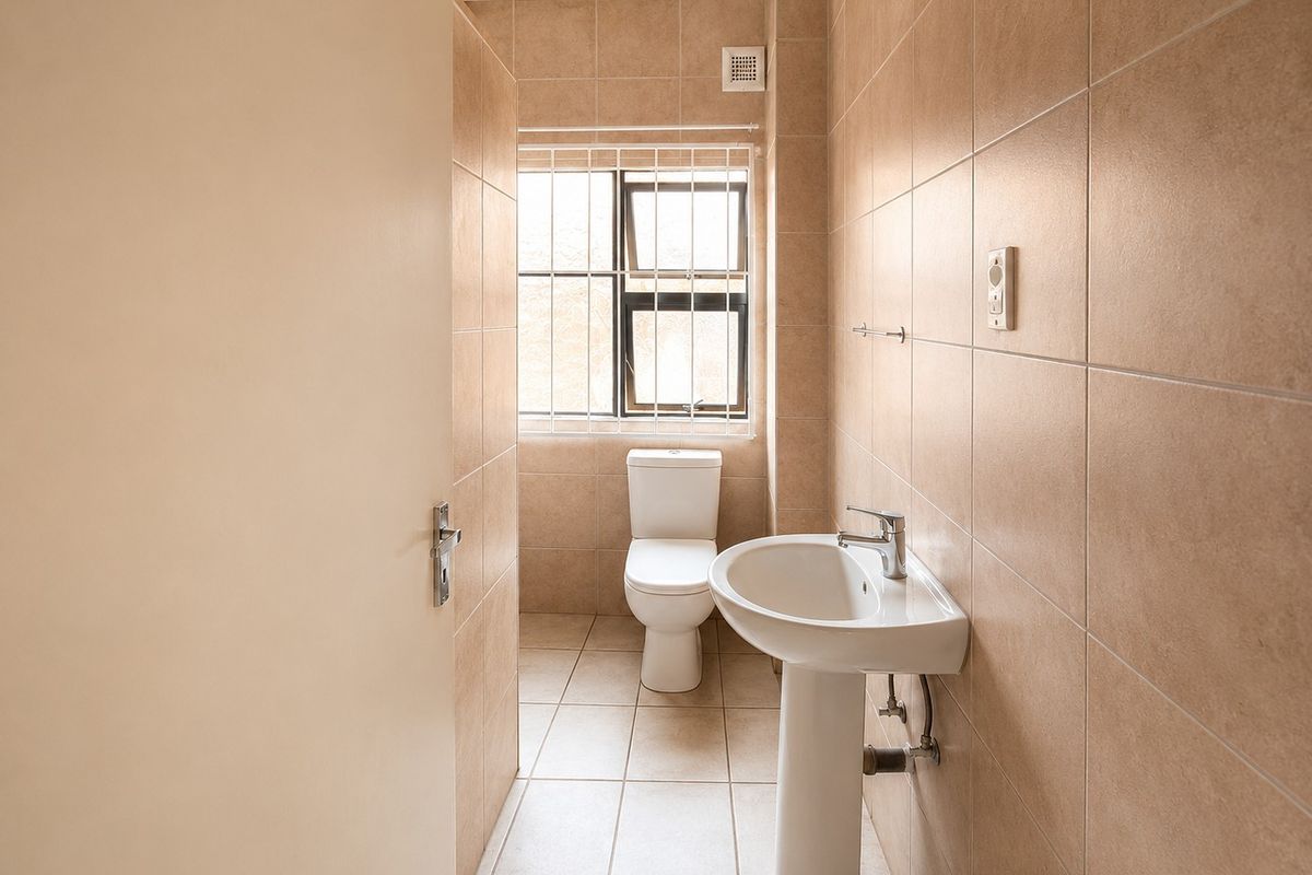 A clean, functional bathroom featuring neutral floor-to-ceiling wall tiles, a pedestal basin, and a toilet, with natural light from a window with security bars.