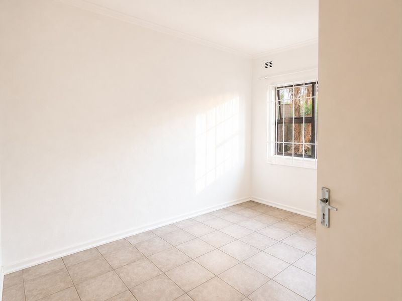 A bright, empty bedroom featuring neutral tiled flooring, clean white walls, and a window fitted with security bars.