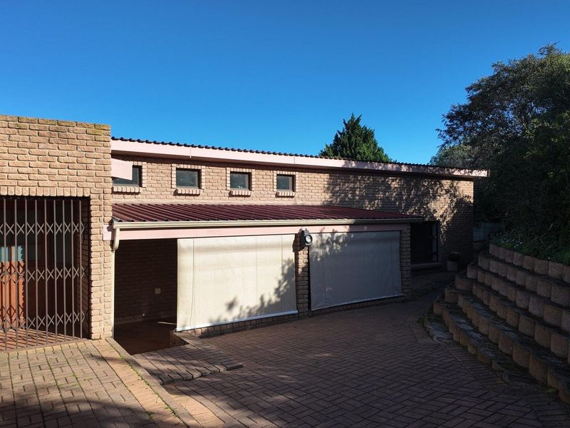 Front exterior view of a brick house featuring a paved driveway, retaining wall, and covered patio area with installed outdoor roller blinds.