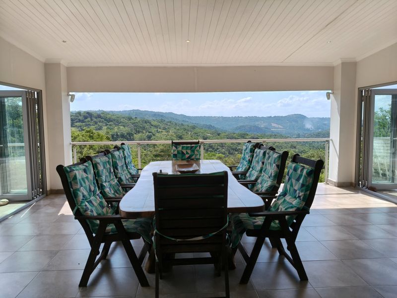 A spacious covered patio featuring tiled flooring, recessed lighting, and a glass balustrade, offering expansive views of the surrounding landscape.