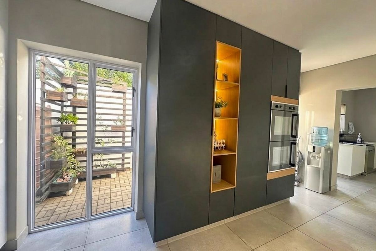 Modern kitchen featuring sleek dark cabinetry, built-in double ovens, integrated shelving, and direct access to an outdoor patio area through a glass door.