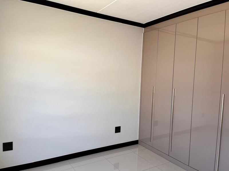 A clean, modern bedroom featuring high-gloss built-in cupboards, neutral tiled flooring, and contrasting black skirting boards.