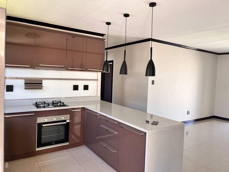 Modern open-plan kitchen featuring high-gloss brown cabinetry, stone countertops, built-in gas hob, oven, and stylish pendant lighting, seamlessly flowing into the living area.