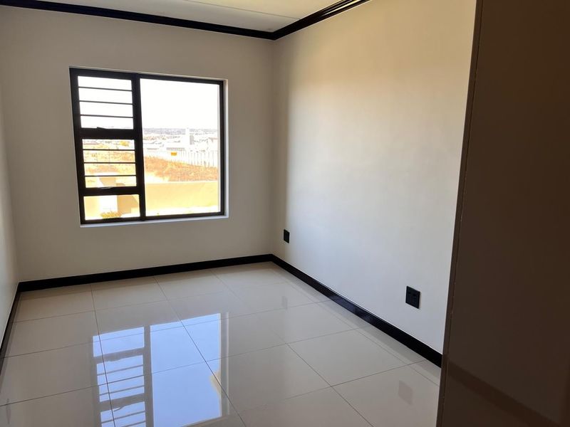 A bright, empty bedroom featuring neutral tiled flooring, clean white walls, and an aluminium-framed window with security bars.