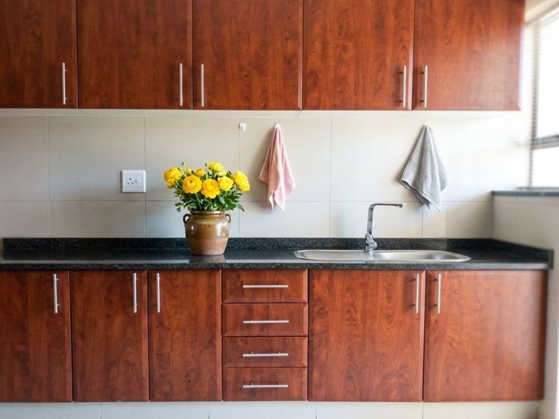 A functional kitchen area featuring wood-finish cabinetry, dark granite countertops, a stainless steel sink, and a neutral tiled backsplash.