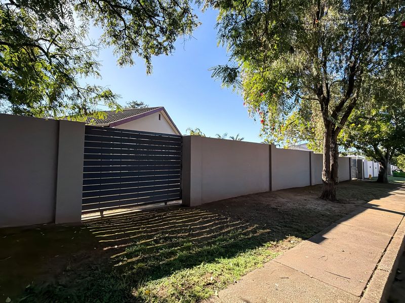 Street view showing the property's perimeter wall, sliding gate, and adjacent sidewalk with mature trees.