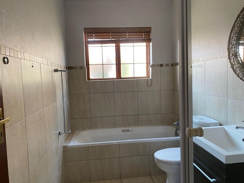 A functional bathroom featuring a built-in bathtub, toilet, and vanity basin with neutral wall tiling and natural light from a window.