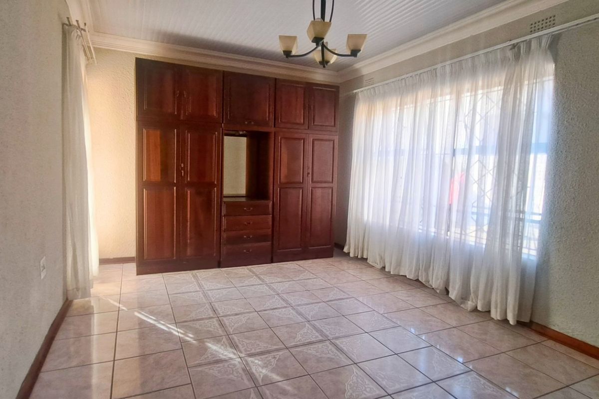 A spacious bedroom featuring built-in wooden cupboards, tiled flooring, and a large window with security bars, offering a clean and functional living space.