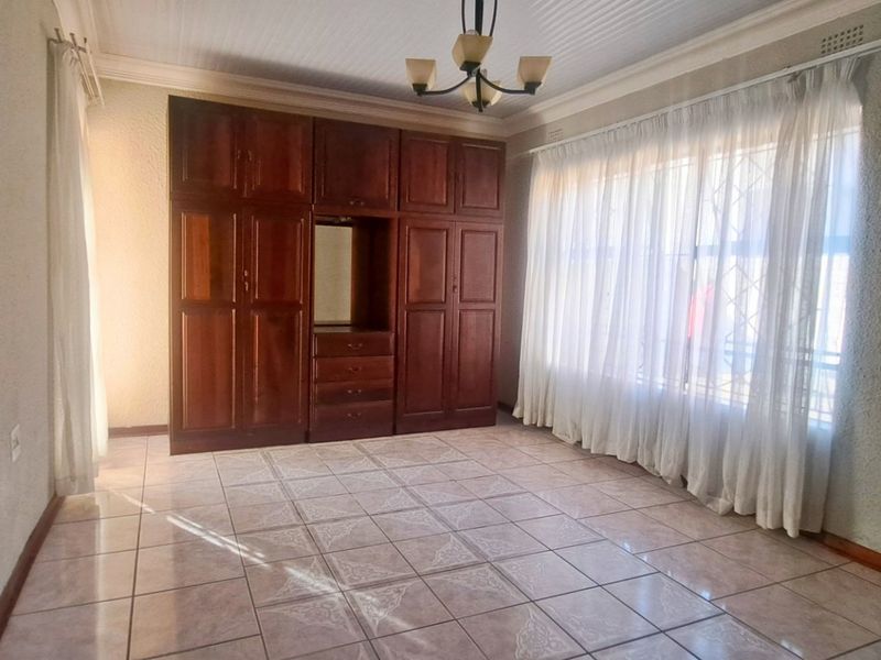 A spacious bedroom featuring built-in wooden cupboards, tiled flooring, and a large window with security bars, offering a clean and functional living space.