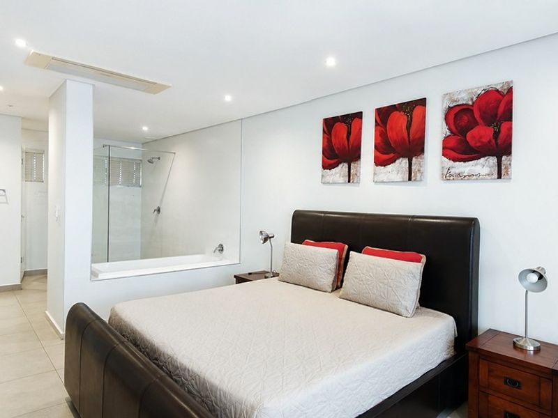 Modern bedroom featuring an open-plan design with an integrated glass-partitioned en-suite bathroom, recessed lighting, and neutral tiled flooring.