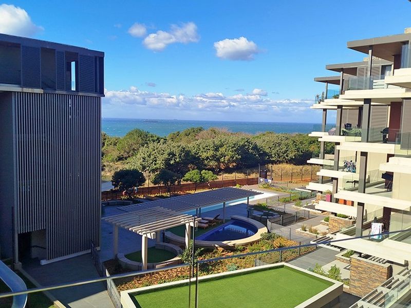 Elevated view overlooking the apartment complex grounds, featuring a swimming pool, landscaped gardens, and a scenic coastal horizon.