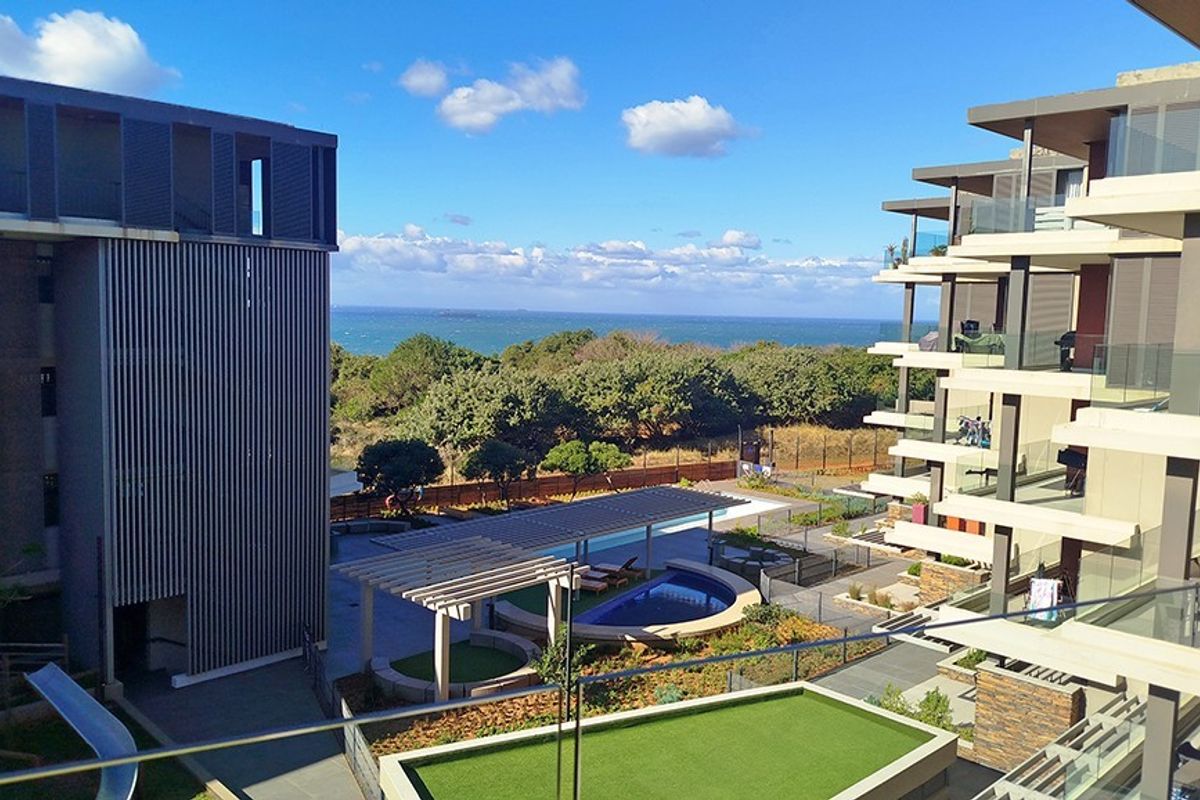 Elevated view overlooking the apartment complex grounds, featuring a swimming pool, landscaped gardens, and a scenic coastal horizon.