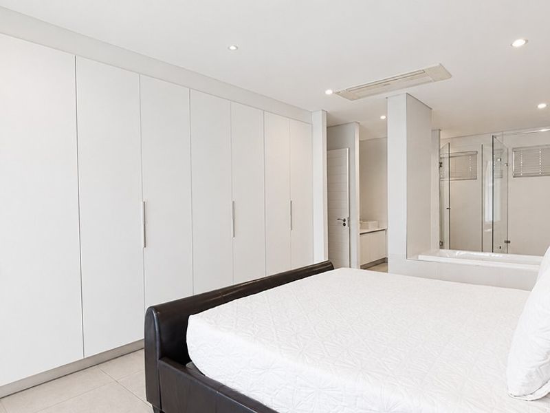 Spacious bedroom featuring extensive built-in cupboards, recessed lighting, ducted air conditioning, and an open-plan en-suite bathroom with a glass shower enclosure.
