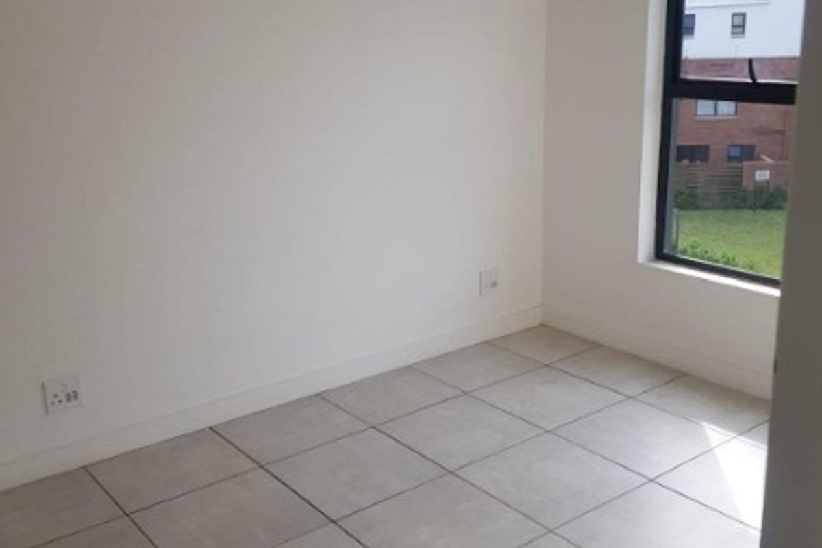 A bright, empty bedroom featuring neutral tiled flooring, a modern pendant light, and a large window with views of the surrounding complex.