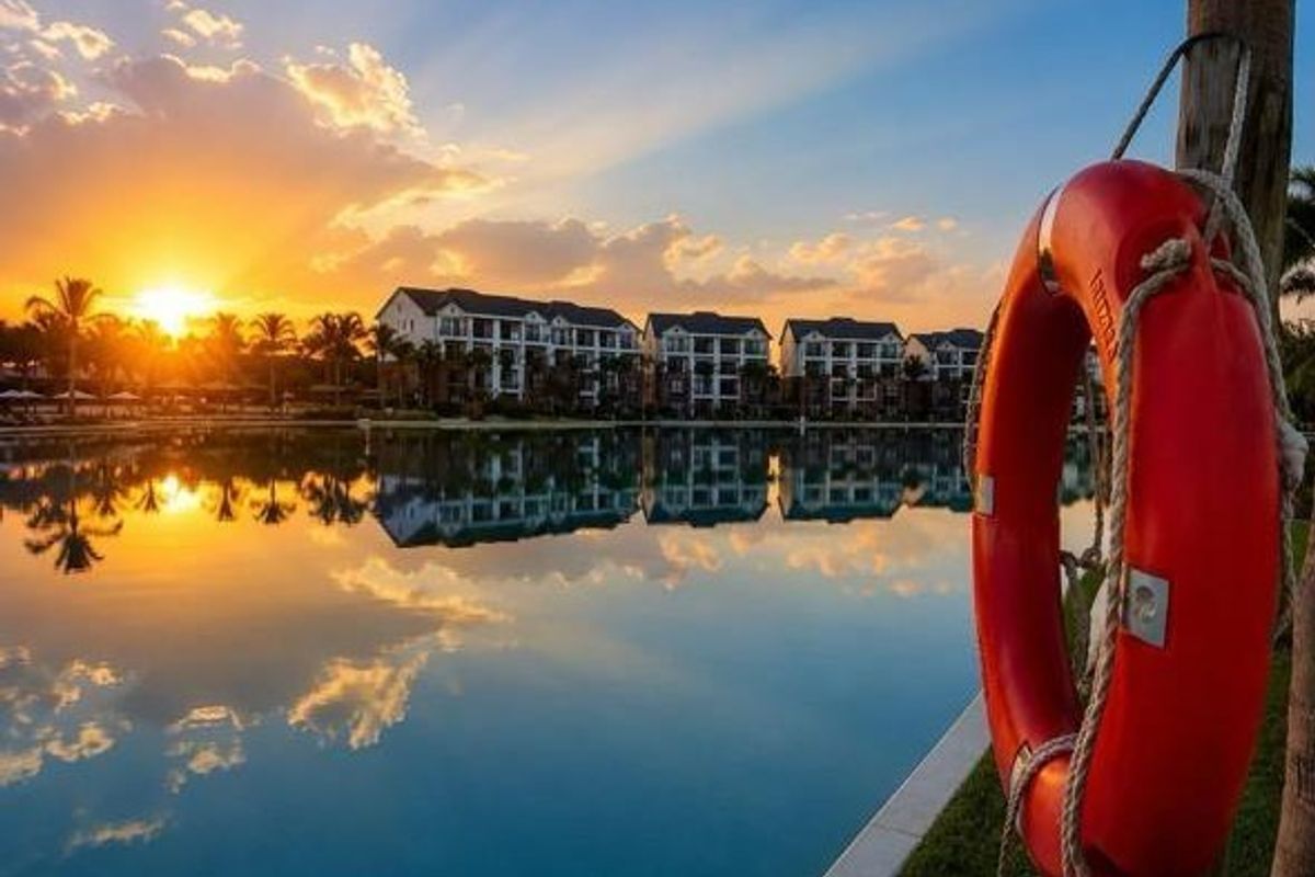 A scenic view of the apartment complex overlooking a calm swimming pool at sunset, featuring a prominent lifebuoy in the foreground.