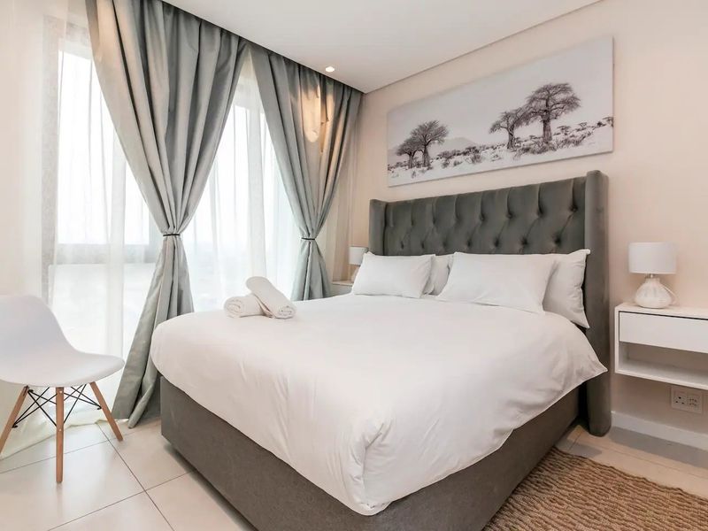 A well-maintained bedroom featuring neutral tiled flooring, recessed ceiling lighting, and floor-to-ceiling curtains providing natural light.