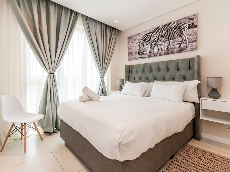 A well-maintained bedroom featuring neutral tones, tiled flooring, recessed lighting, and large windows with floor-to-ceiling curtains.