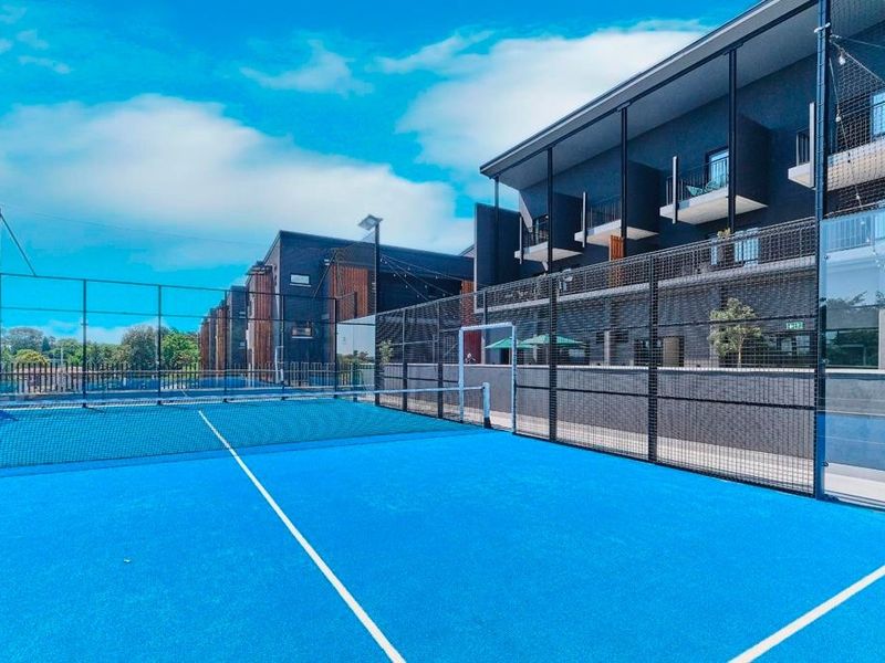 A well-maintained outdoor padel court facility located within an apartment complex, featuring modern fencing, floodlighting, and adjacent residential balconies.