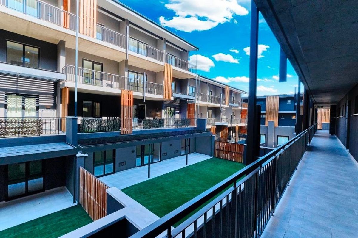 A view of the modern apartment complex exterior, featuring multi-level balconies, metal railings, timber privacy screens, and a well-maintained artificial turf courtyard area.