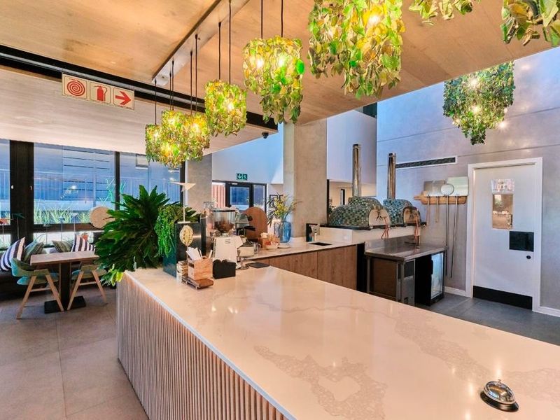 Modern open-plan commercial kitchen and service area featuring stone countertops, timber cabinetry, and professional pizza ovens in a well-lit, contemporary space.