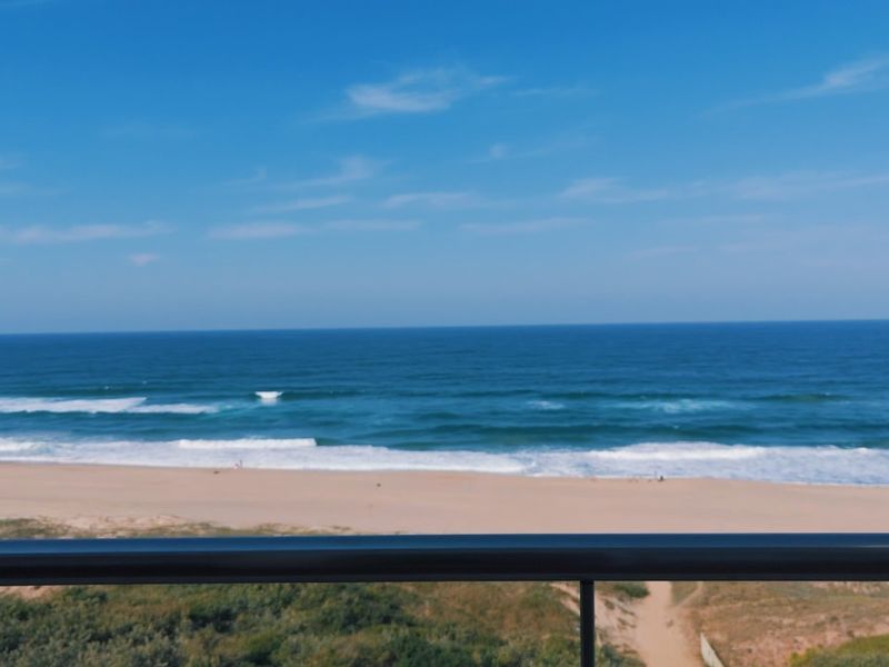 A clear, elevated view of the ocean and sandy beach, captured from a balcony with a modern railing.