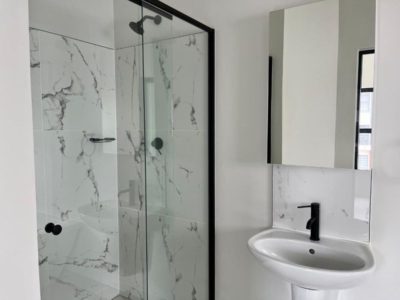 Modern bathroom featuring a walk-in shower with black-framed glass, marble-look wall tiles, a pedestal sink with a black faucet, and a wall-mounted mirror.